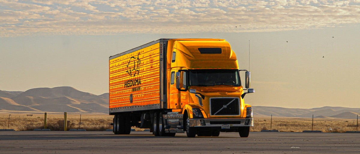 Yellow truck with a logo on the side driving on a road with mountains in the background